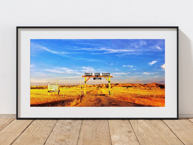 May include: A panoramic view of a rural landscape with a wooden sign that reads "Circle View Ranch" above a dirt road leading into a field of golden grass. The sky is blue with white clouds.