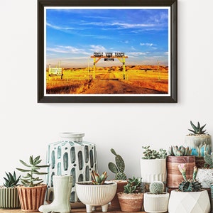 May include: A framed photograph of a rustic wooden gate with the words "Circle View Ranch" on a sign above it. The gate is open and leads to a dirt track that disappears into the distance. The background is a field of golden grass under a bright blue sky with fluffy white clouds.
