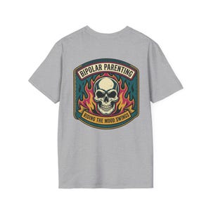 May include: A gray t-shirt featuring a graphic with a skull, flames, and text that reads "Bipolar Parenting Riding the Mood Swings." The design is in a vintage style with a color palette of teal, yellow, and red.