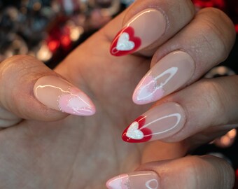 May include: Close-up of a hand with long, almond-shaped nails painted with a pink and white base. Each nail features a white heart design with a red outline.