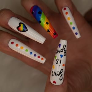 Può includere: Unghie lunghe bianche con disegni arcobaleno. Un'unghia ha un cuore arcobaleno, un'altra ha punti arcobaleno e un'altra ha le parole "Love is!" con piccoli cuori.