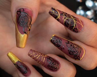 May include: Close-up of five long, almond-shaped nails with a burgundy base and gold accents. Each nail features a black rose design with intricate details.