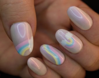 Puede incluir: Primer plano de cinco uñas ovaladas pintadas con una base rosa claro. Tres uñas presentan un diseño de arcoíris con nubes blancas, mientras que las otras dos uñas son de color rosa claro sólido.