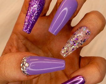 Op de afbeelding: Lange, valse nagels in paars en glitter. De nagels zijn in verschillende stijlen, waaronder effen paars, glitter en helder met glitter.
