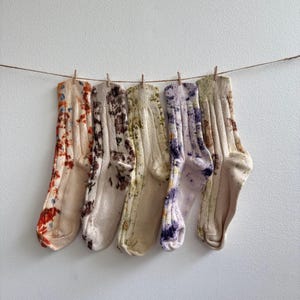 May include: Five pairs of ribbed crew socks hanging on a thin string. Each pair features a unique botanical-inspired tie-dye pattern in shades of orange, brown, green, purple, and tan. The socks have a textured knit finish and ribbed cuffs.