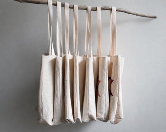 May include: A collection of six off-white canvas tote bags with long shoulder straps, all hanging from a single horizontal piece of weathered driftwood. The bags feature a simple rectangular shape and a textured fabric surface.