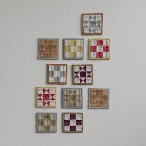 May include: A collection of ten square quilted coasters featuring various geometric patterns and colours including shades of brown, grey, yellow, red, and purple. Each coaster has a stitched fabric border and visible quilting lines throughout.