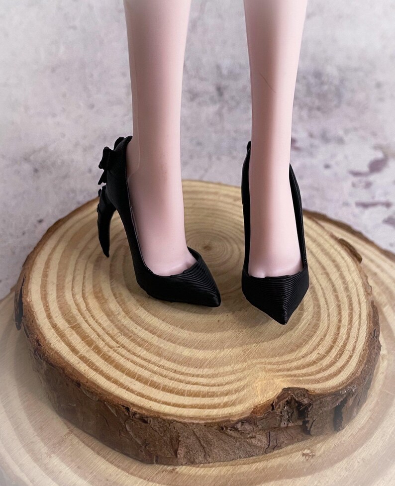 以下が含まれることがあります： ポインテッドトゥとヒールにリボンディテールが付いた黒いハイヒール。靴は木製の表面にあります。ヒールは約7.6cmです。