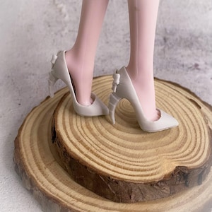 May include: A pair of off-white high heels with pointed toes and bow details on the heels. The shoes are on a wooden surface, with the legs of a person wearing the shoes visible.