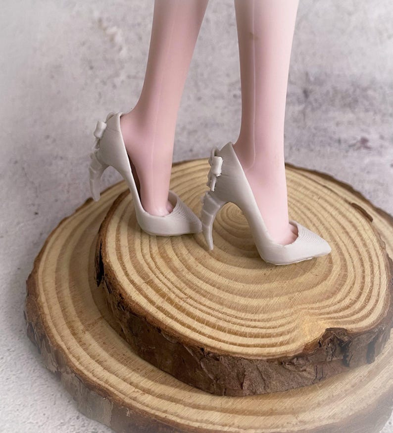 May include: A pair of off-white high heels with pointed toes and bow details on the heels. The shoes are on a wooden surface, with the legs of a person wearing the shoes visible.
