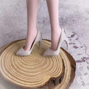 May include: A pair of light gray high-heeled shoes with a unique design. The heels have a curved shape with decorative elements. The shoes are on a wooden surface, showcasing the footwear's details and style.