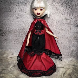 Könnte beinhalten: Eine Puppe mit weißen Haaren, die einen roten Umhang und ein schwarz-rotes Kleid mit Spitzenbesatz trägt. Die Puppe hat rote Augen und eine schwarze Halskette.