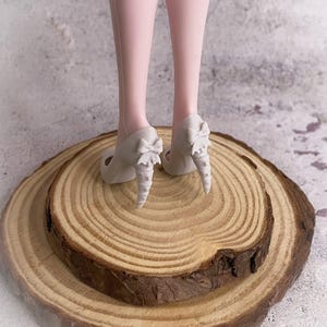 May include: A pair of light gray high heels with bow accents on the heels. The heels are placed on a wooden slice, with the legs of a doll visible above the shoes. The background is a neutral, textured surface.