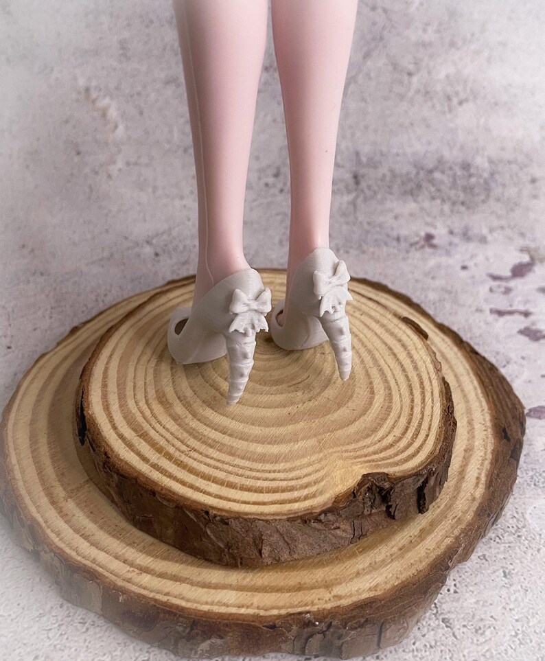 May include: A pair of light gray high heels with bow accents on the heels. The heels are placed on a wooden slice, with the legs of a doll visible above the shoes. The background is a neutral, textured surface.