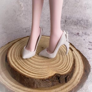 May include: A pair of light gray high-heeled shoes with pointed toes and decorative accents on the heels. The shoes are displayed on a wooden slice with visible growth rings. The heels appear to be approximately 3 inches tall.