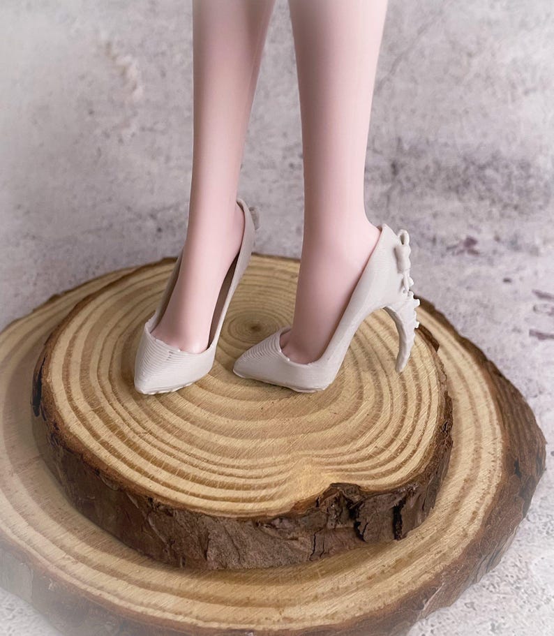 May include: A pair of light gray high-heeled shoes with pointed toes and decorative accents on the heels. The shoes are displayed on a wooden slice with visible growth rings. The heels appear to be approximately 3 inches tall.