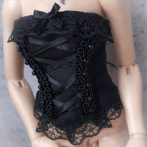 Op de afbeelding: Zwart korset met kanten afwerking en lintdetails. De bustier heeft een veter aan de voorkant en is versierd met kleine, donkere kralen. Een kleine strik zit in het midden bovenaan. Het korset is ontworpen voor een pop.