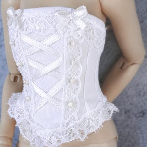 May include: White corset with lace trim, bows, and pearl accents. The bodice features a criss-cross ribbon design and small pearl-like beads. The corset is designed for a doll or similar small figure.