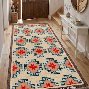 May include: Rectangular beige rug with a geometric pattern in red, blue, and peach. The rug features a repeating design of star-like shapes and angular lines, with a brown border and fringe.