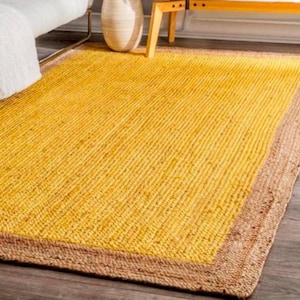 Puede incluir: Una alfombra rectangular con una textura tejida. El centro es de color amarillo brillante, enmarcado por un borde marrón natural. La alfombra está colocada sobre un suelo de madera, con un vistazo a los muebles en el fondo.