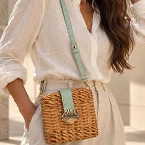 Peut inclure: Un sac bandoulière carré en rotin tressé doté d'une sangle texturée vert clair et d'une patte de fermeture verticale assortie. Le sac est orné d'un fermoir en métal doré en forme de coquillage centré sur le rabat avant.