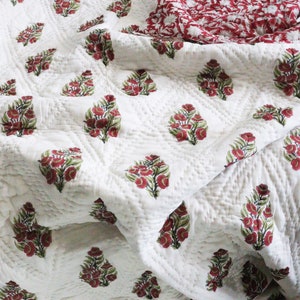 May include: White cotton quilt with a repeating pattern of red and green floral designs. The quilt is folded and has a textured surface.