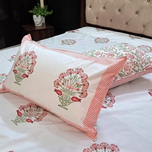 Könnte beinhalten: Weiße Bettwäsche mit einem floralen Muster in Rosa und Grün. Zwei rechteckige Kissenbezüge mit passendem floralen Design und gestreiftem Rand liegen auf dem Bett. Auch die Bettdecke weist das florale Muster auf.