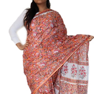 May include: A traditional Indian saree with a floral print in shades of orange, red, and white. The saree is made of cotton and has a border with a repeating pattern.