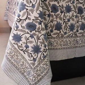 Op de afbeelding: Een wit tafelkleed met een blauw en grijs bloemenpatroon. Het tafelkleed heeft een rand met een gestreept patroon.