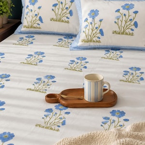 Puede incluir: Una sábana blanca y dos fundas de almohada a juego con un patrón floral azul repetitivo y tallos verdes. Una bandeja de madera sostiene una taza de cerámica a rayas azules y blancas y una cuchara dorada, junto a una manta beige texturizada.