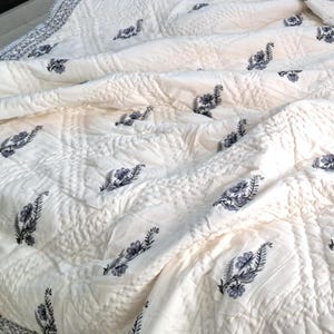 Puede incluir: Una colcha de algod&oacute;n blanco con un sutil patr&oacute;n acolchado y un delicado dise&ntilde;o floral en tonos grises. La colcha est&aacute; doblada y drapeada sobre una cama.