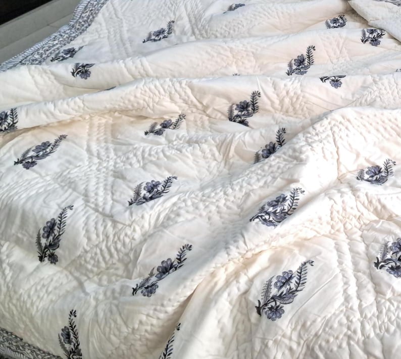 Puede incluir: Una colcha de algod&oacute;n blanco con un sutil patr&oacute;n acolchado y un delicado dise&ntilde;o floral en tonos grises. La colcha est&aacute; doblada y drapeada sobre una cama.