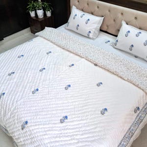May include: White quilted bedspread and pillow set with a blue floral pattern. The bedding features a repeating design of blue flowers and leaves. The pillows and the border of the bedspread have a striped pattern.