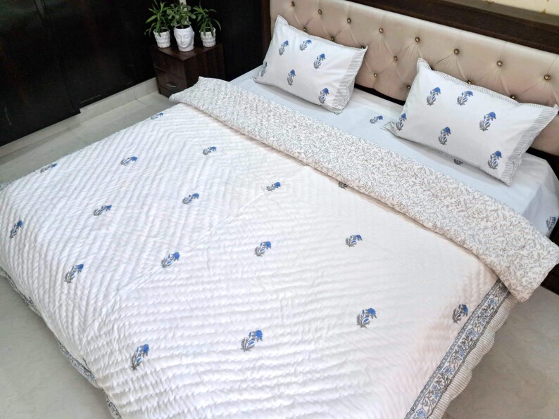 May include: White quilted bedspread and pillow set with a blue floral pattern. The bedding features a repeating design of blue flowers and leaves. The pillows and the border of the bedspread have a striped pattern.