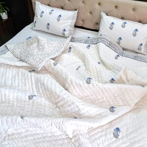 May include: White quilted bedspread with blue floral accents and matching pillowcases. The bedding features a striped border and a decorative pillow with a floral pattern. The bed is made up with a tufted headboard.