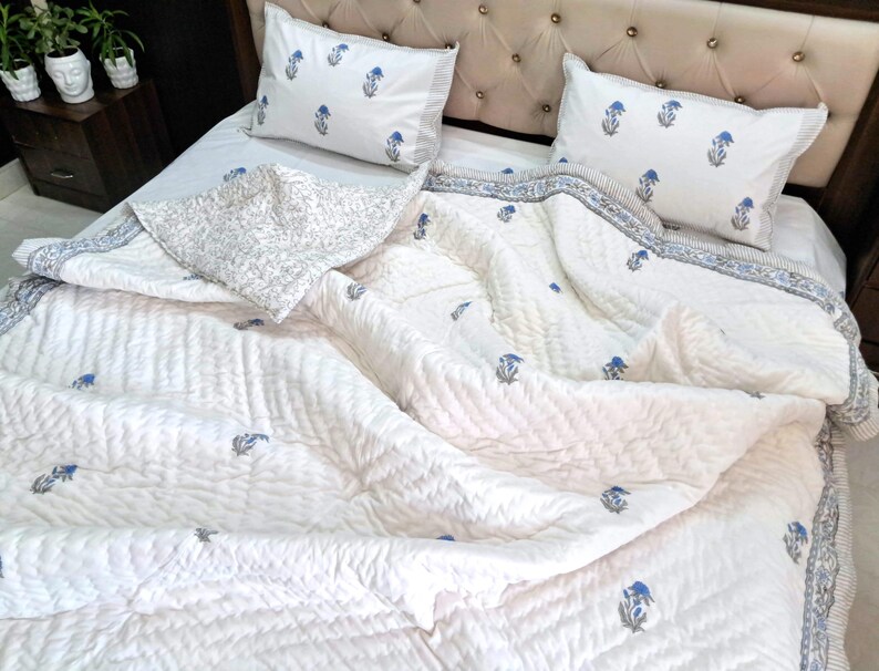 May include: White quilted bedspread with blue floral accents and matching pillowcases. The bedding features a striped border and a decorative pillow with a floral pattern. The bed is made up with a tufted headboard.