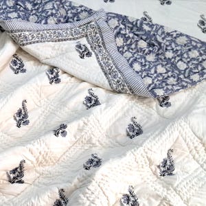 Puede incluir: Una colcha de algodón blanco con un estampado floral azul. La colcha está doblada por la mitad y tiene una textura acolchada.