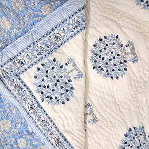 Op de afbeelding: Een wit katoenen dekbed met een blauw bloemenpatroon. Het dekbed heeft een rand van blauwe en witte strepen. Het dekbed is dubbelgevouwen en het bloemenpatroon is aan beide kanten zichtbaar.