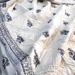 Puede incluir: Una colcha de algod&oacute;n blanco con estampado floral azul. La colcha tiene un borde con un patr&oacute;n a rayas.