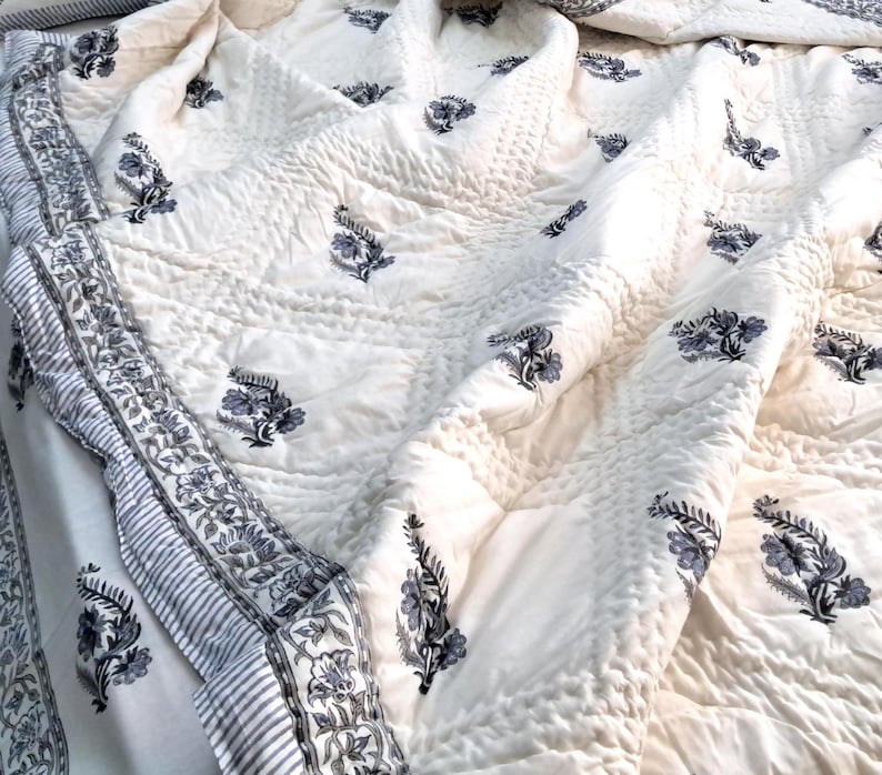 Puede incluir: Una colcha de algod&oacute;n blanco con estampado floral azul. La colcha tiene un borde con un patr&oacute;n a rayas.