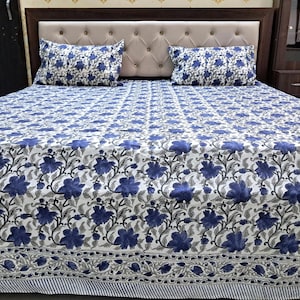 Puede incluir: Un edredón blanco y azul con estampado floral y dos fundas de almohada a juego. La ropa de cama presenta un patrón repetido de flores azules y hojas grises sobre un fondo blanco. El edredón tiene un borde a rayas.