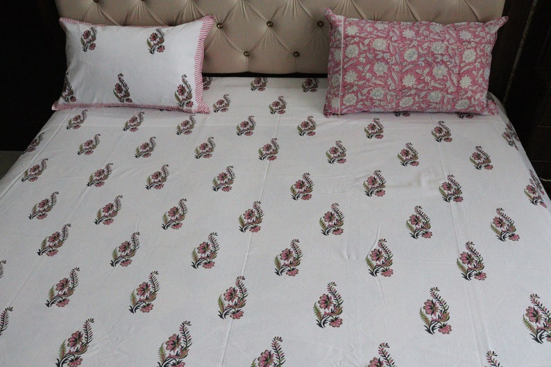 May include: White cotton bed sheet with a pink floral pattern. The sheet is on a bed with two matching pillowcases, one with a pink floral pattern and the other with a white floral pattern.