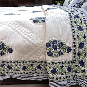May include: A white quilted bedspread with a floral pattern in navy blue and green. The bedspread has a striped border and is folded over, revealing the pattern and texture. The overall theme is bedding.