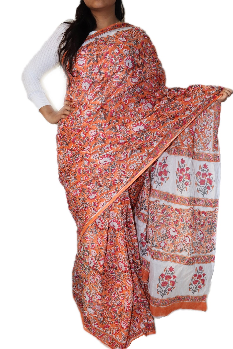 May include: A traditional Indian saree with a floral print in shades of orange, red, and white. The saree is made of cotton and has a border with a repeating pattern.