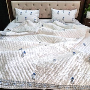 May include: White quilted bedding set with blue floral accents. The set includes a comforter and two matching pillowcases. The comforter features a geometric pattern and a blue floral border. The pillows also have the same blue floral design.