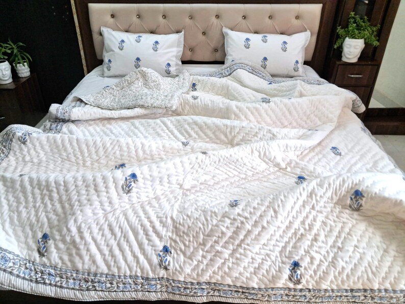 May include: White quilted bedding set with blue floral accents. The set includes a comforter and two matching pillowcases. The comforter features a geometric pattern and a blue floral border. The pillows also have the same blue floral design.