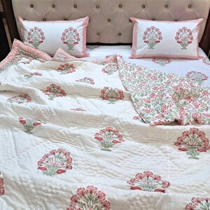 May include: A white quilted bedspread and two matching pillowcases with a floral pattern in red and green. The bedding set is on a bed with a beige headboard.