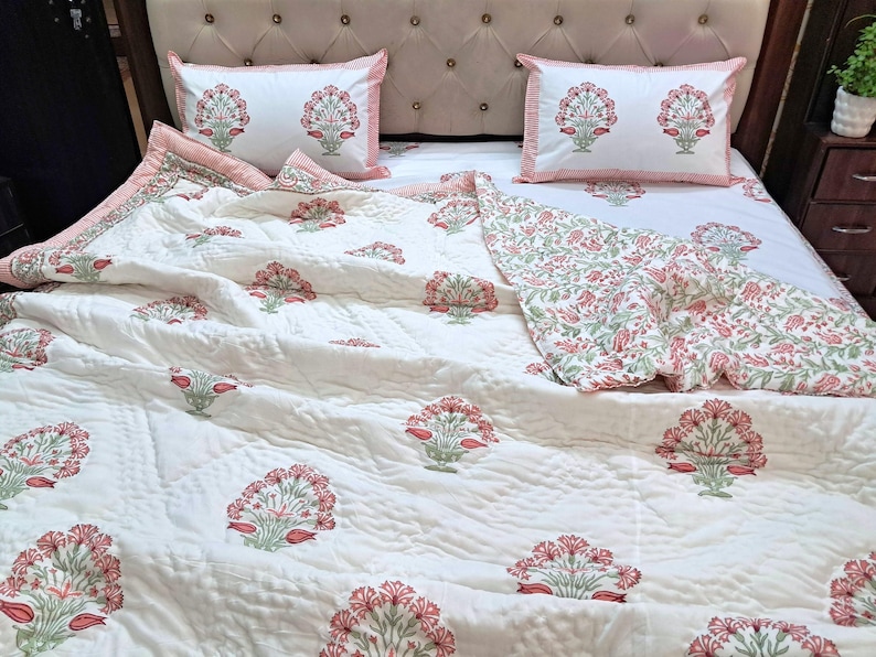 May include: A white quilted bedspread and two matching pillowcases with a floral pattern in red and green. The bedding set is on a bed with a beige headboard.