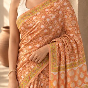 May include: A peach-colored saree featuring an all-over white floral print, accented by a yellow and orange border with intricate botanical patterns. The garment is draped over a simple, sleeveless cream-colored blouse, showcasing a traditional design.