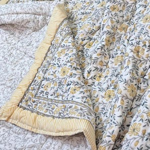 May include: A quilted blanket with a floral pattern in yellow and gray on a white background. The border is a yellow and white striped design. The blanket is folded, showing the pattern and texture of the fabric.